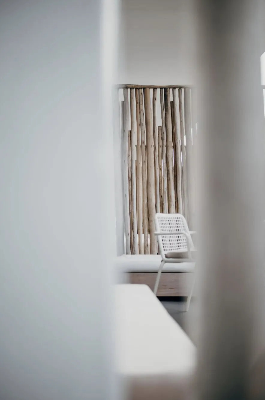 A glimpse between soft white walls of a quiet room: a screen of pale driftwood poles, a white woven chair and a low bench.