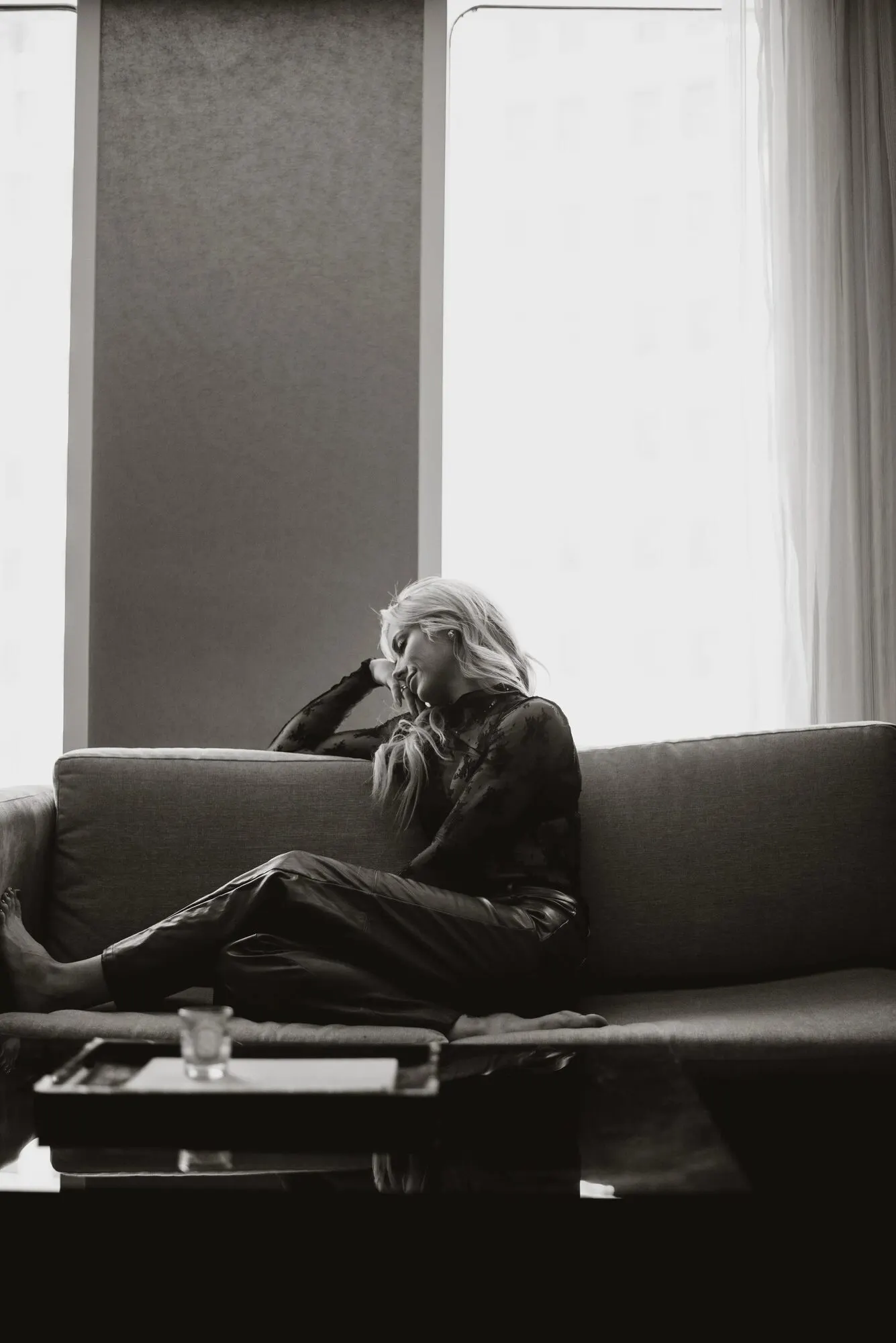 Figure seated low on a couch in a high-floor room, city light coming through the glass.