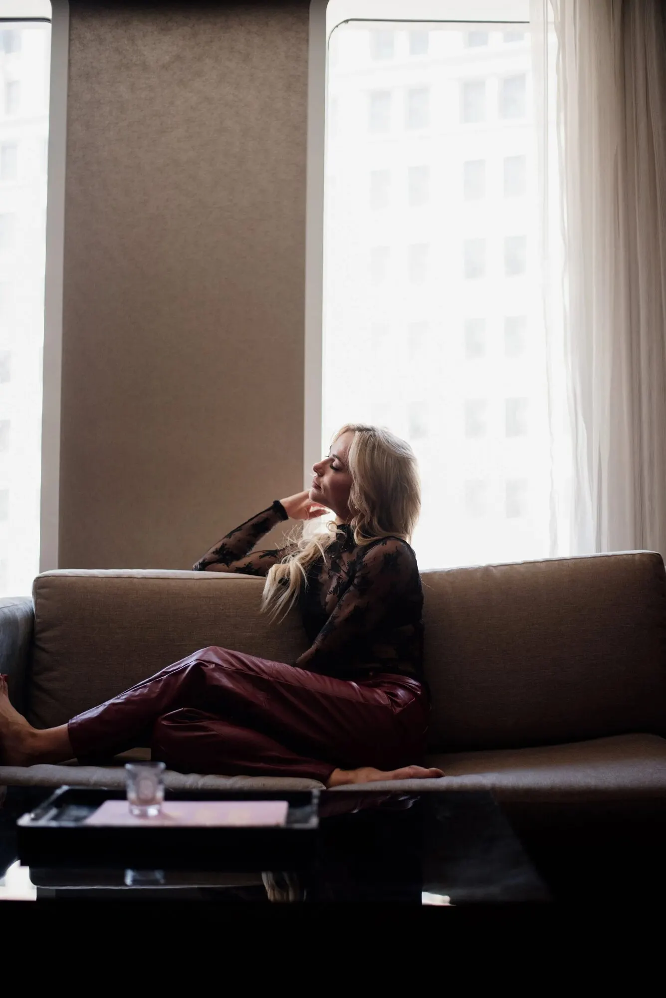 Tyler Tanner sitting back on a low hotel couch in oxblood leather trousers, city towers filling the windows behind her.