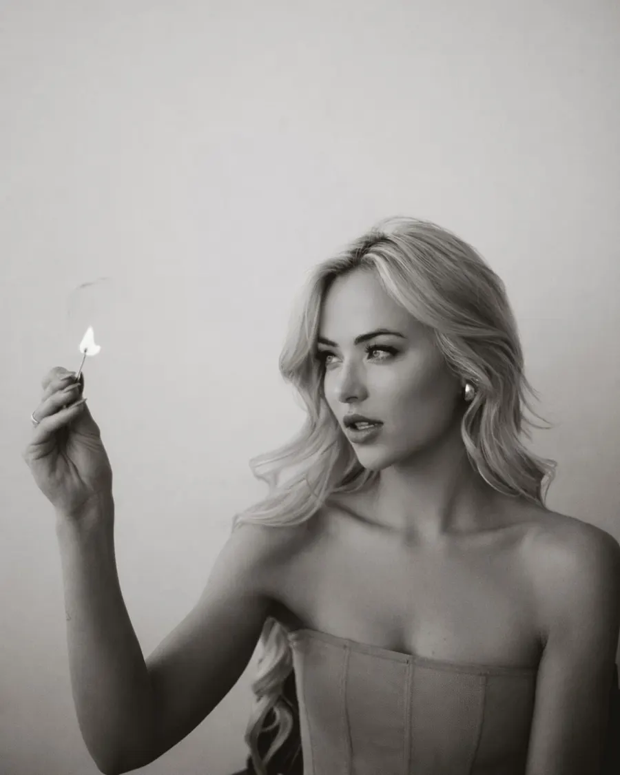Tyler Tanner holding a lit match, black and white, corseted, the flame the brightest thing in frame.
