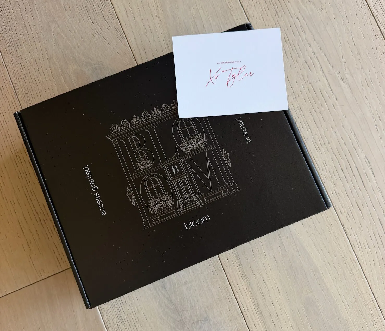 A black Bloom presentation box on pale timber floor with a handwritten note card resting on top.